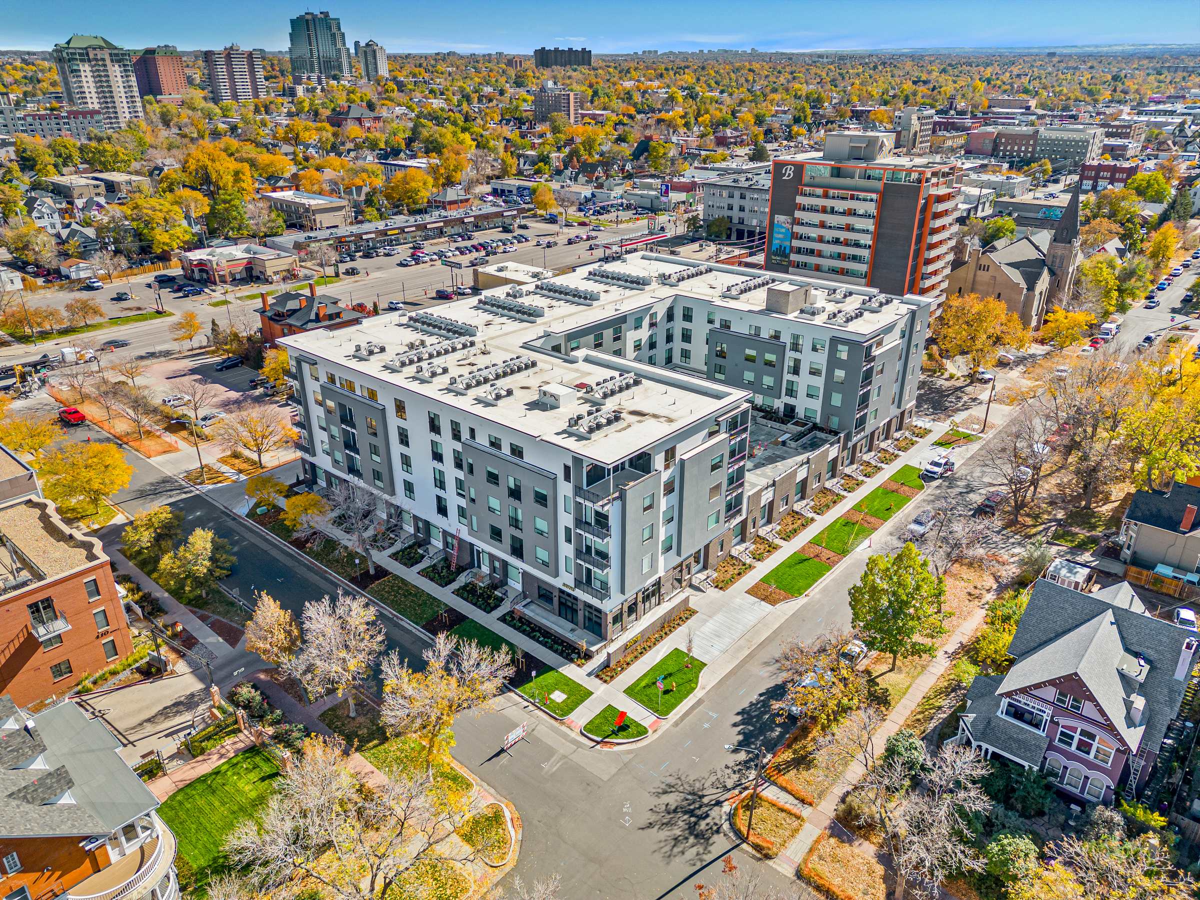 Aerial view of a modern apartment complex in a city neighborhood with trees, cars, and surrounding buildings.