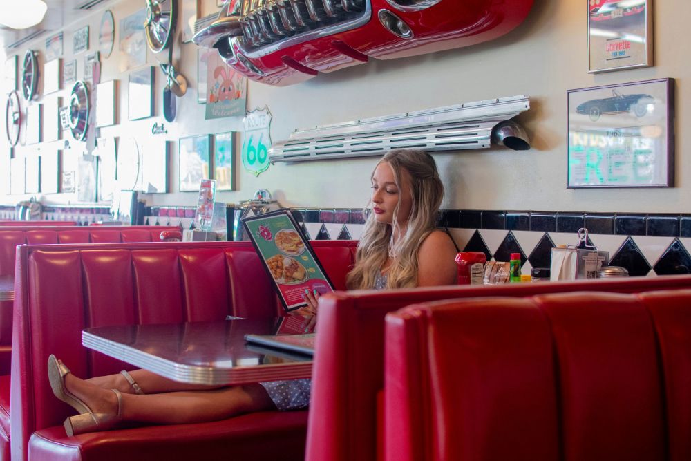A woman sits alone in a retro diner booth reading a menu, surrounded by vintage decor and red seats.